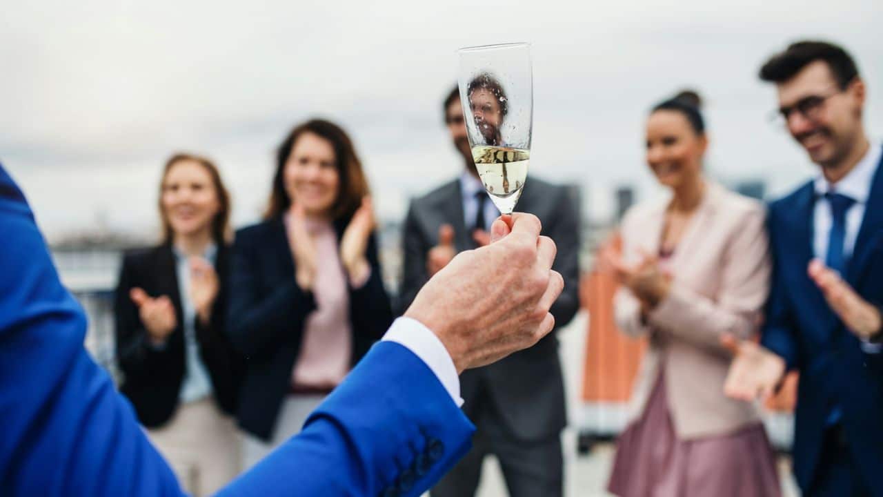 A person in a blue suit raises a glass of champagne to a group of applauding people.