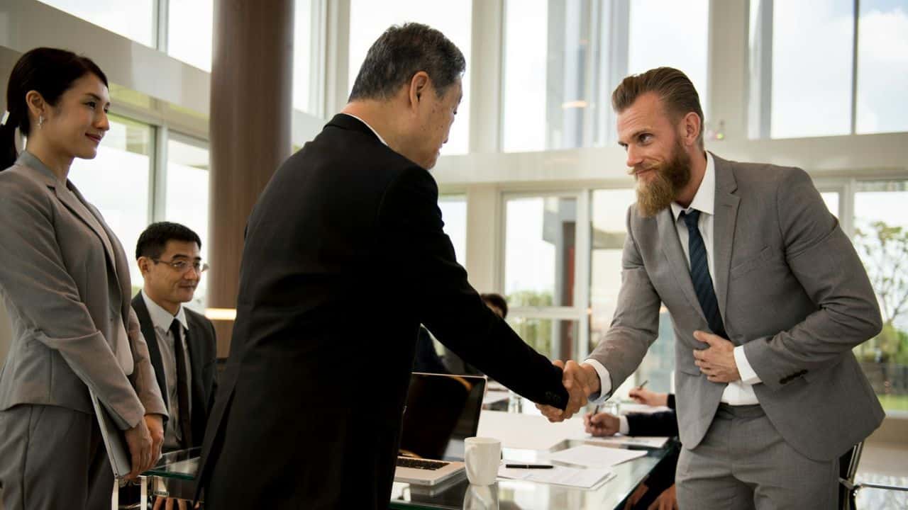 A bearded man in a suit shakes hands with another man in a business setting.