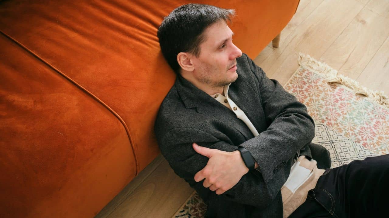 A man in a dark jacket sits on a wooden floor, leaning against an orange sofa.