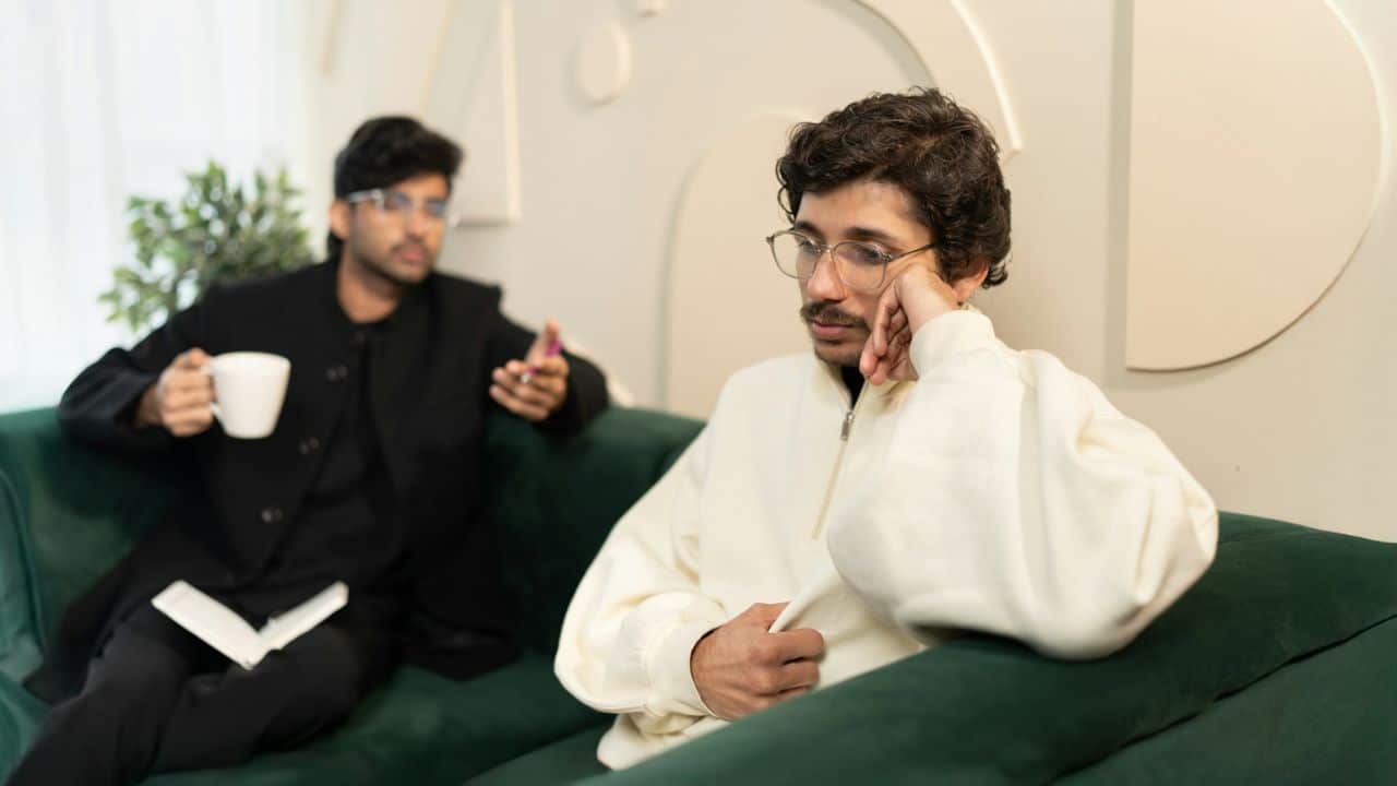 A man in a cream jacket with his hand on his chin looks thoughtful, while another man gestures.