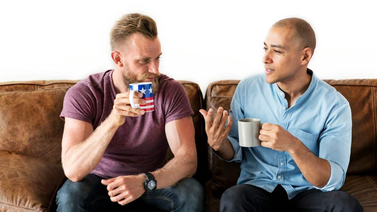 Two men sit on a brown couch, drinking from mugs and engaged in conversation.