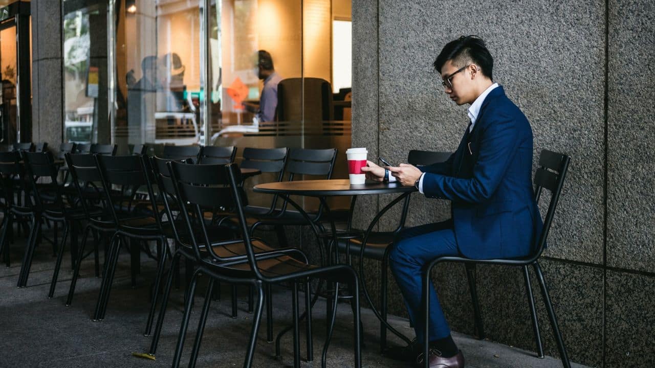 A man in a blue suit sits at an outdoor cafe table, looking at his phone.