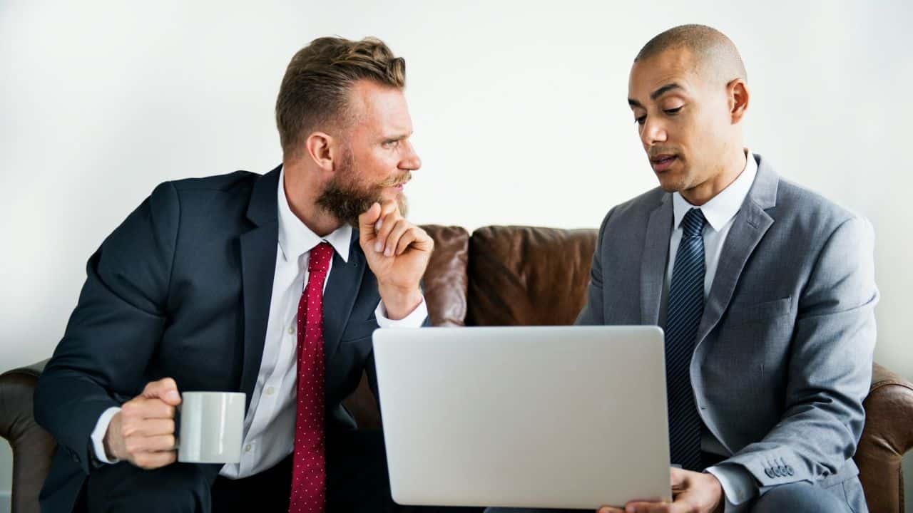 Two men in suits sit on a couch with a laptop, one holding a mug and the other gesturing.
