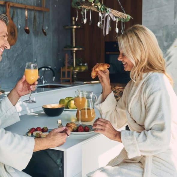 A man and woman in white bathrobes are smiling at each other while having breakfast.