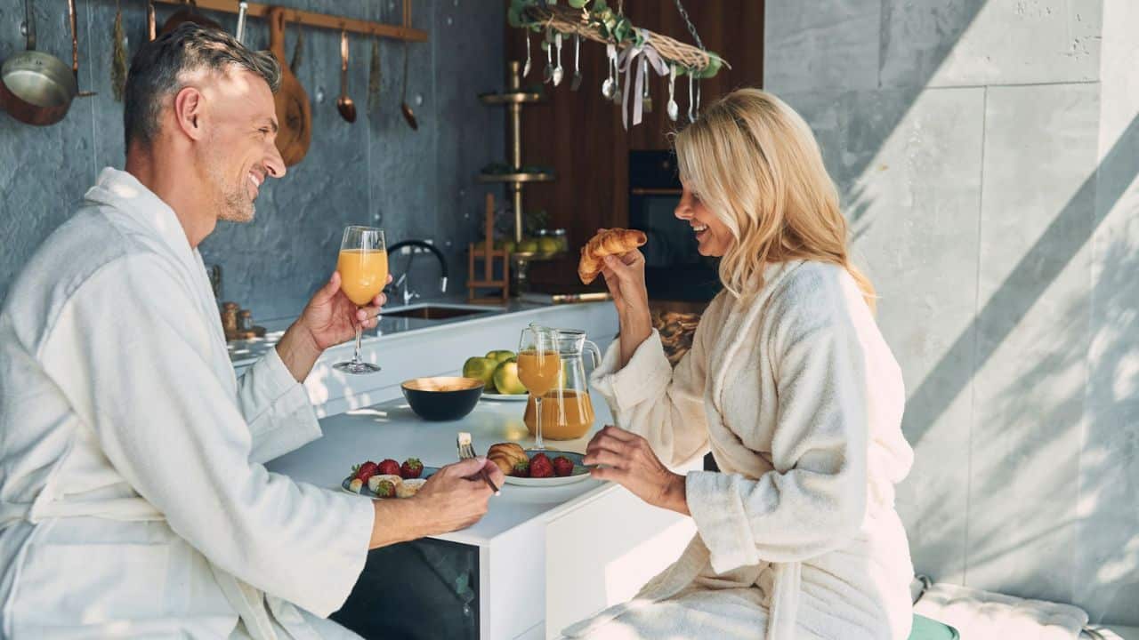 A man and woman in white bathrobes are smiling at each other while having breakfast.