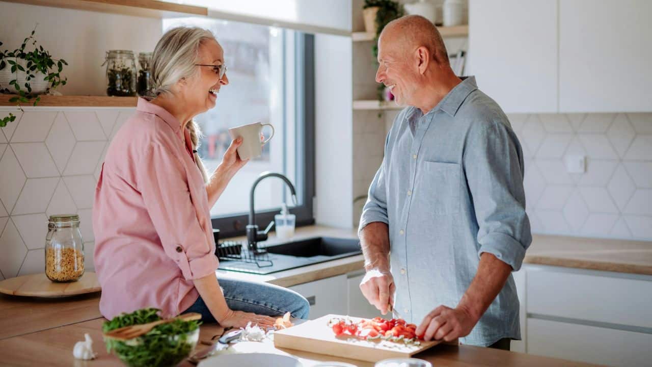 A man chops vegetables while a woman sits on the counter, both smiling in a kitchen.