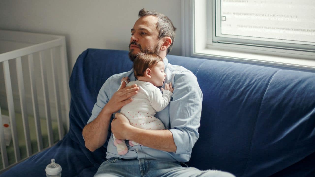 A man sits on a blue couch, holding a sleeping baby in his arms.