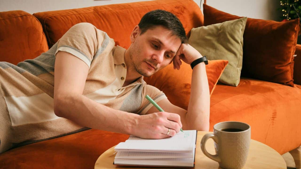 A man lies on an orange sofa, writing in a notebook with a pen, next to a coffee mug.