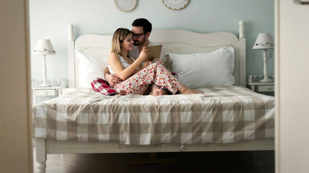A happy couple sits in bed, smiling and looking at a tablet together.