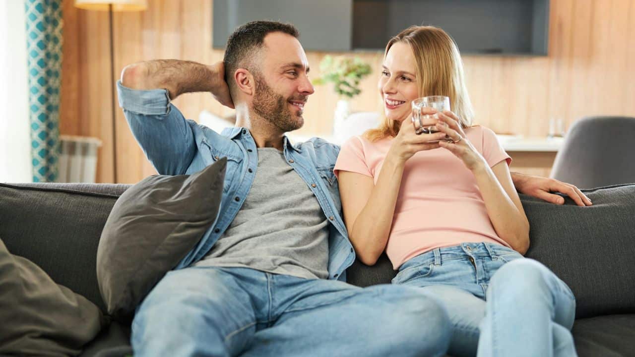 A couple smiles at each other while relaxing on a sofa, the woman holding a glass.