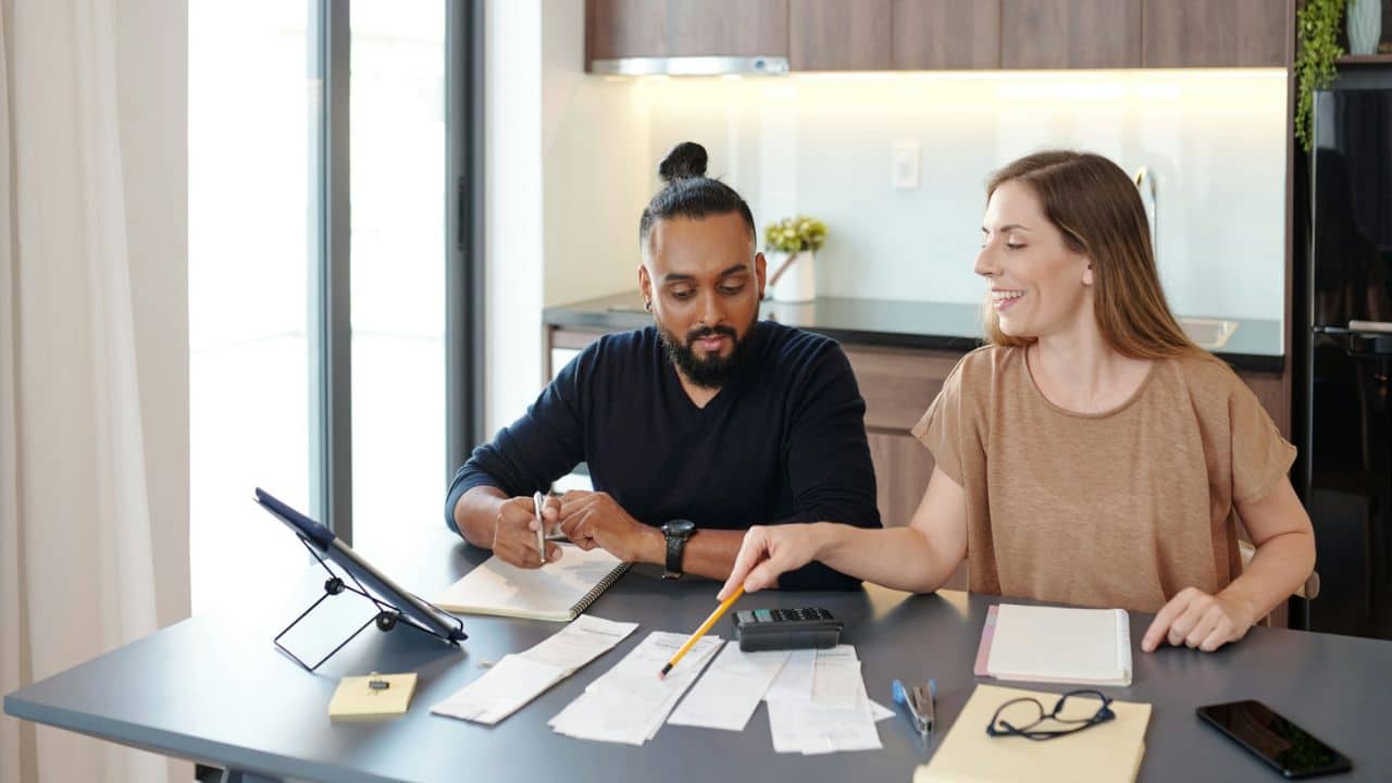 A couple sits at a table, reviewing papers and writing, with a tablet and calculator nearby.