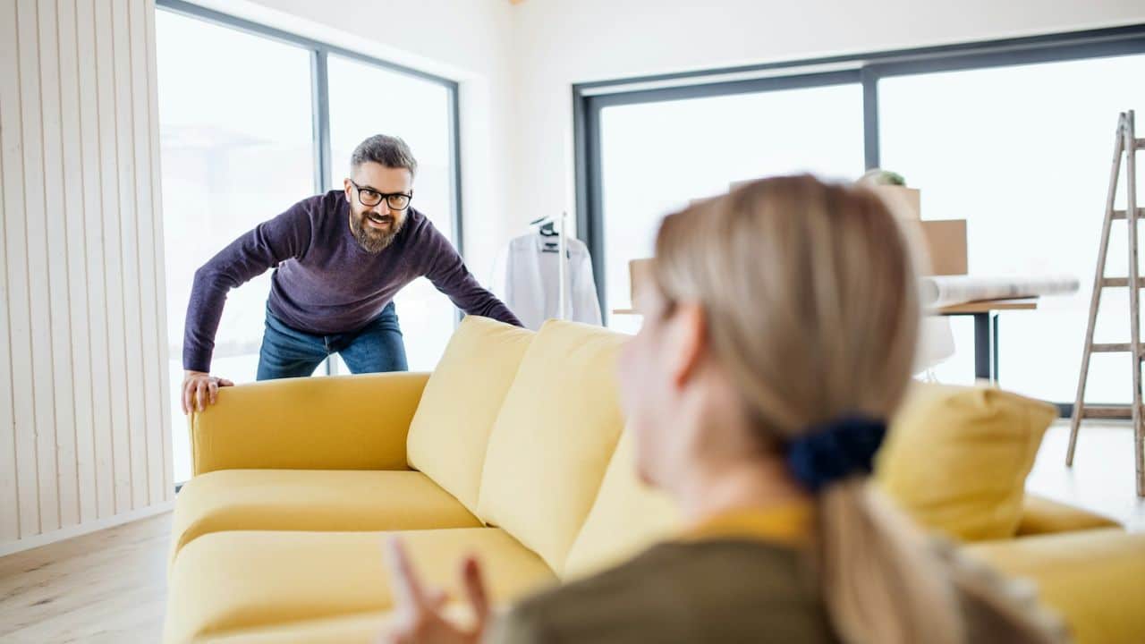 A man pushes a yellow sofa while a woman sits in the foreground, facing away.