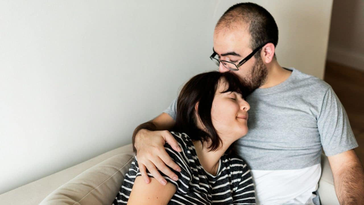A man wearing glasses gently kisses a woman on the forehead, both sitting on a sofa.