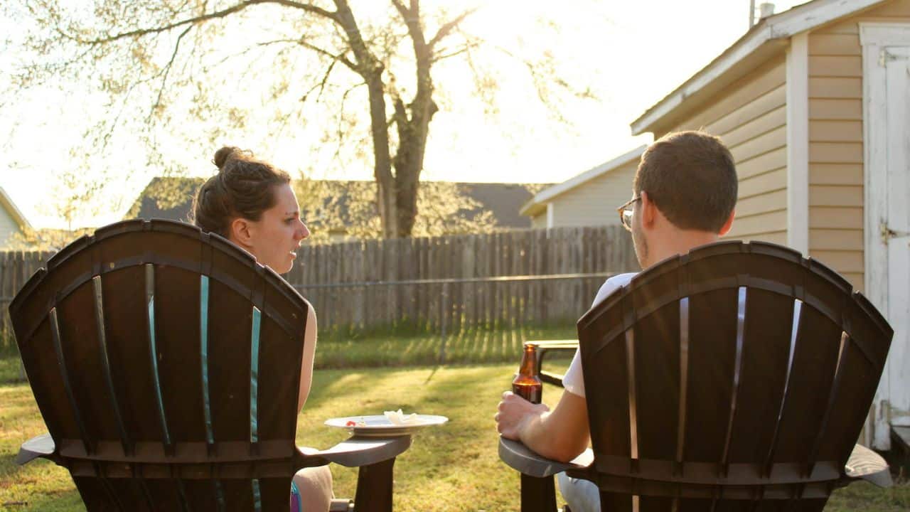 A man and a woman sit in Adirondack chairs outdoors, facing each other at sunset.
