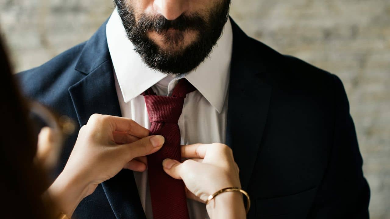 A person's hands adjust a man's red tie on his navy suit and white shirt.