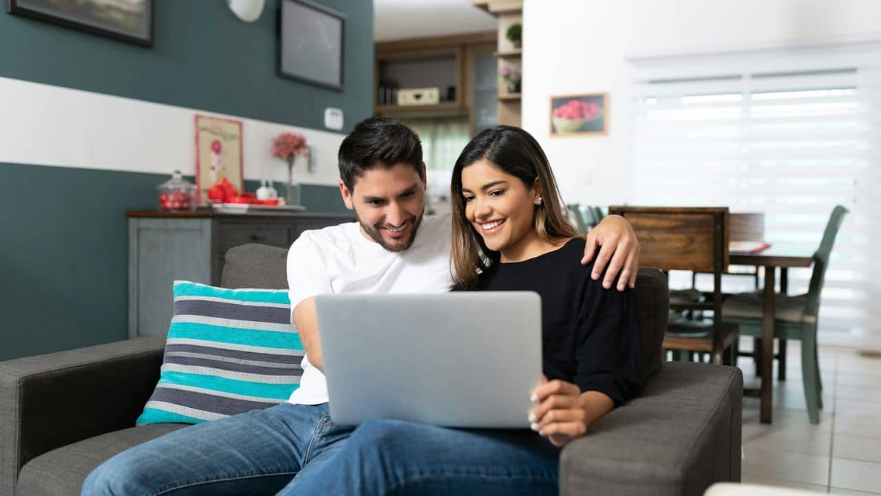 A smiling couple sits on a sofa, looking at a laptop together.