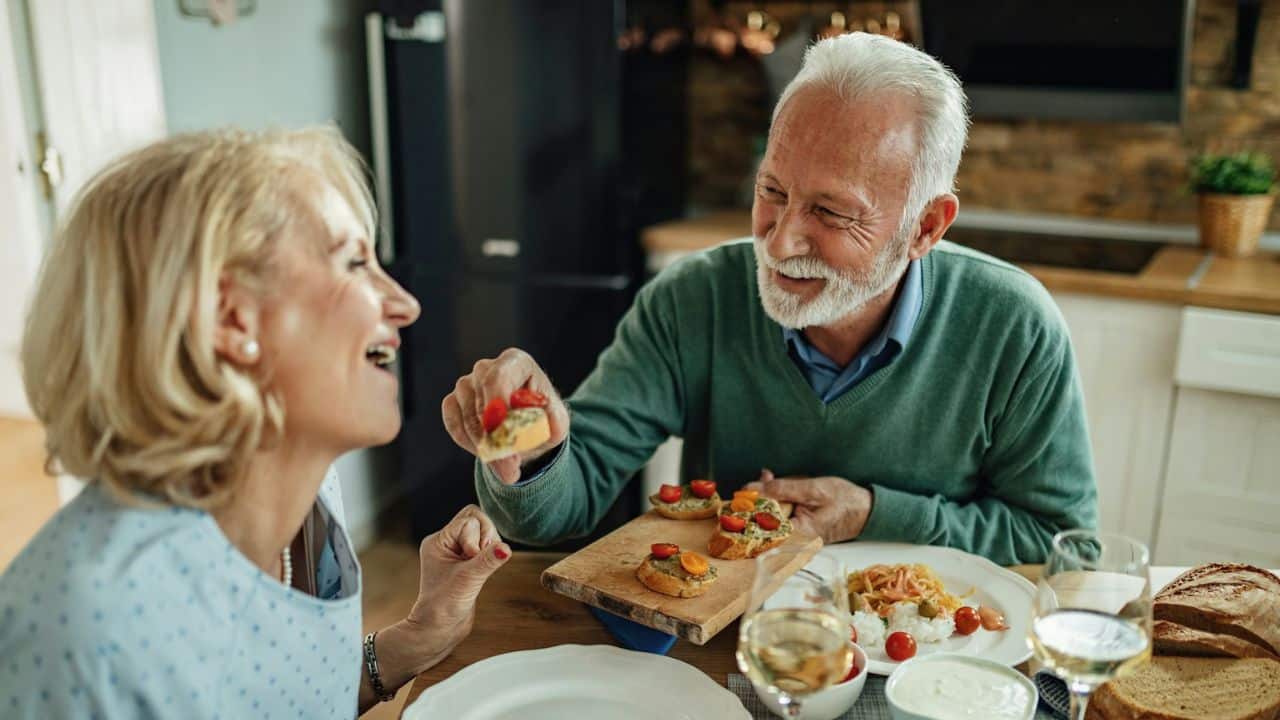 A smiling man with a beard offers a piece of bruschetta to a laughing woman at a dining table.