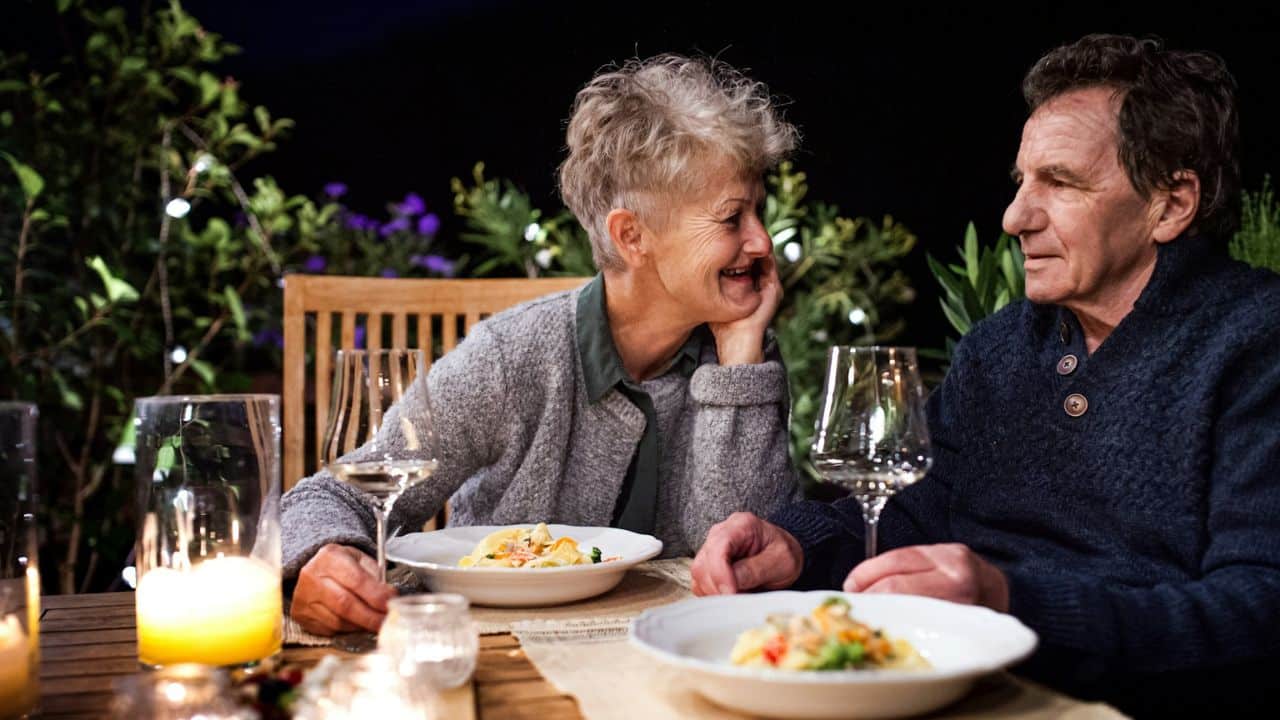 A smiling older woman looks at a man across a candlelit table with food and wine glasses.