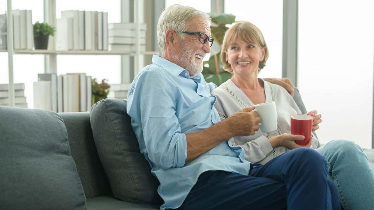 A smiling older couple sits on a couch, holding mugs, looking at each other.