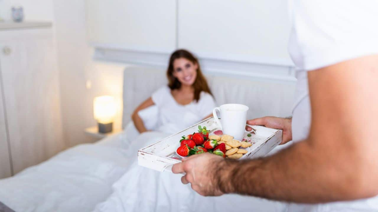 A person brings a breakfast tray with strawberries and cookies to a smiling woman in bed.