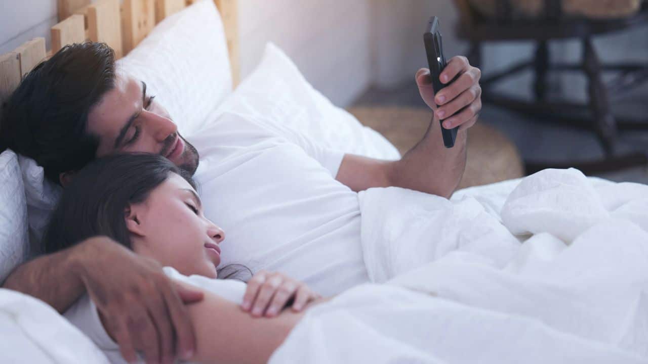A man in bed holds a phone, while a woman sleeps with her head on his arm.