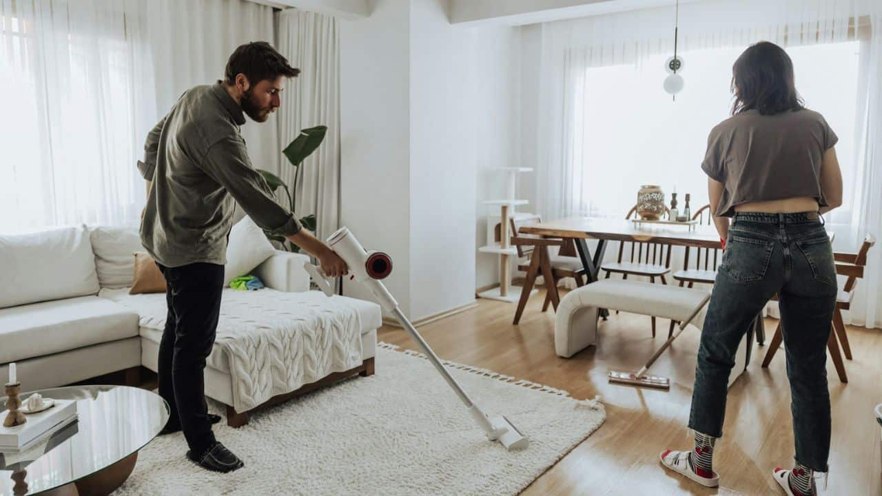 A man vacuums a rug while a woman stands nearby in a living and dining area.