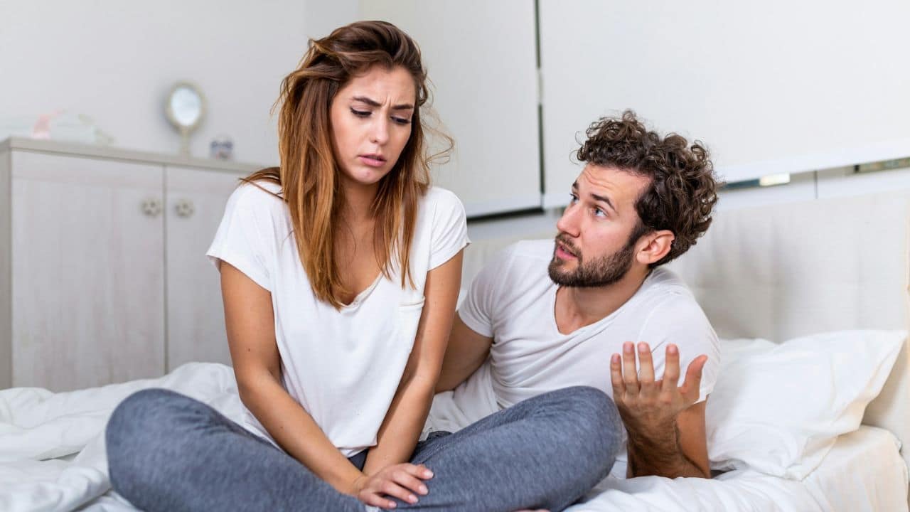 A man gestures while talking to a woman who looks upset, both in bed.