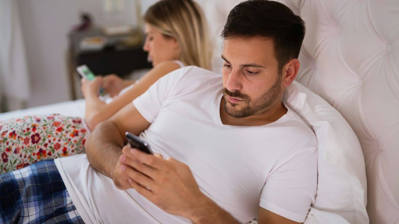 A man and woman lie in bed, both looking at their individual phones.
