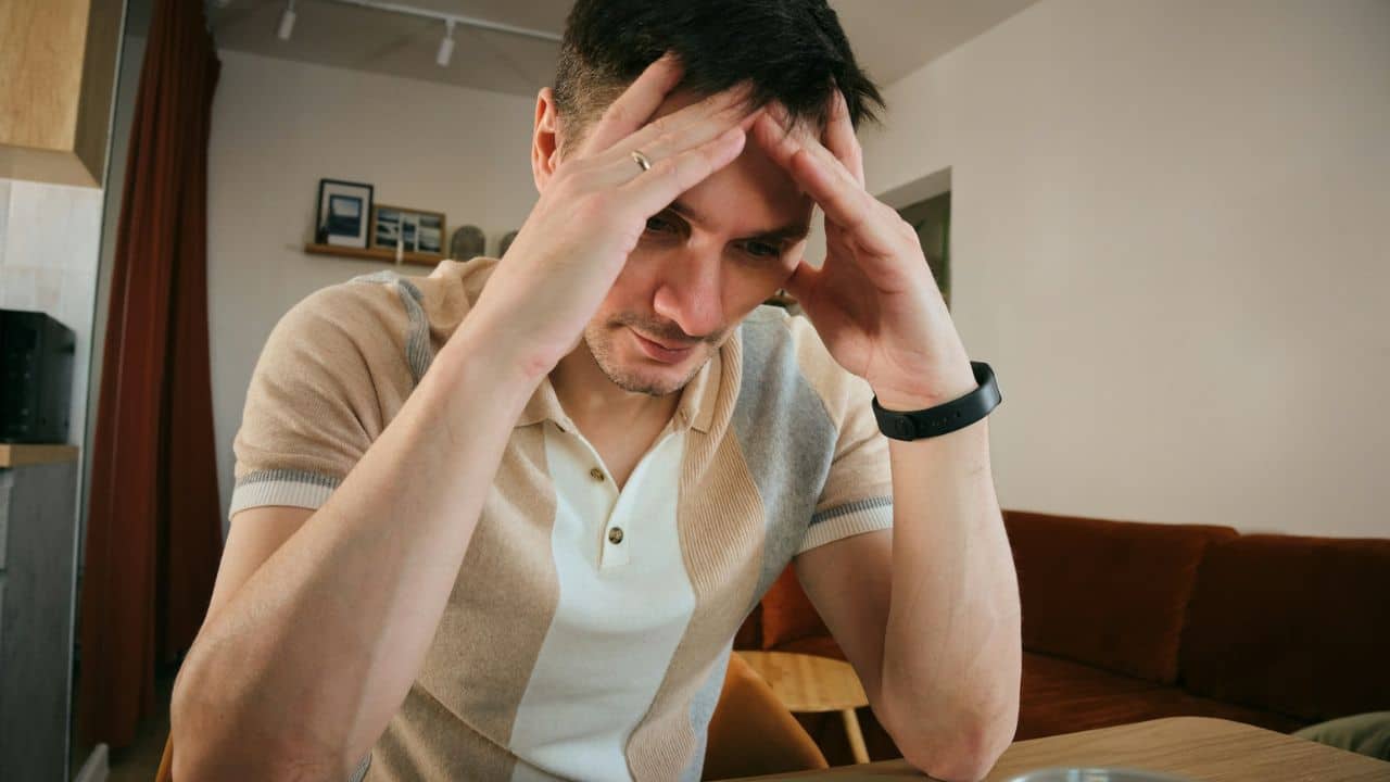 A man with short hair sits, holding his head, looking stressed or tired.