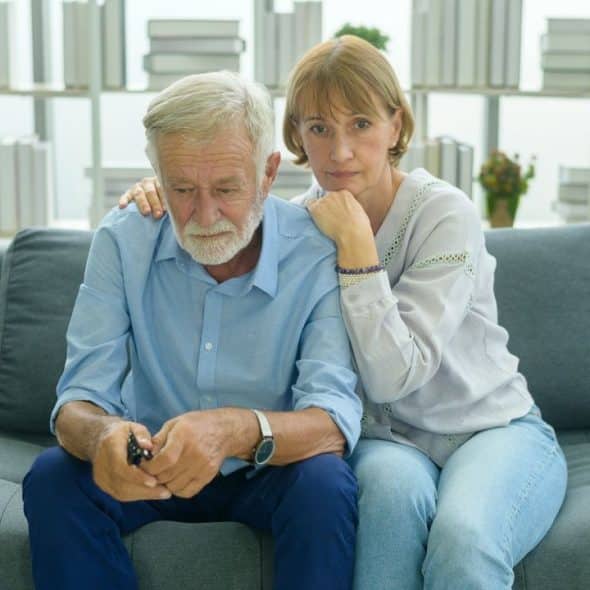 An older man sits on a couch, looking down, while a woman rests her hand on his shoulder.