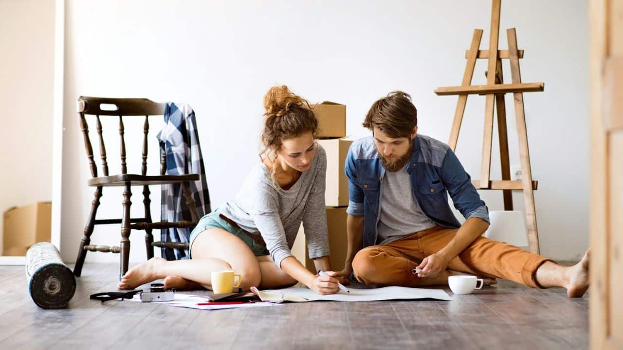A couple sits on a wooden floor, looking at papers, surrounded by moving boxes and art supplies.