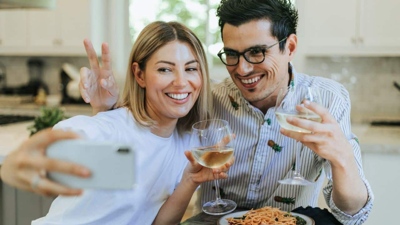 A happy couple takes a selfie, holding wine glasses, with food on the table.