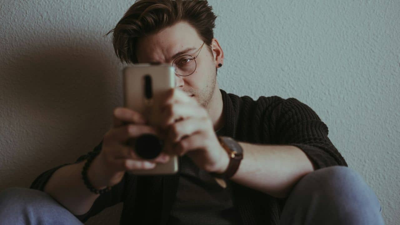 A man wearing glasses and a dark sweater sits against a wall, holding a smartphone.