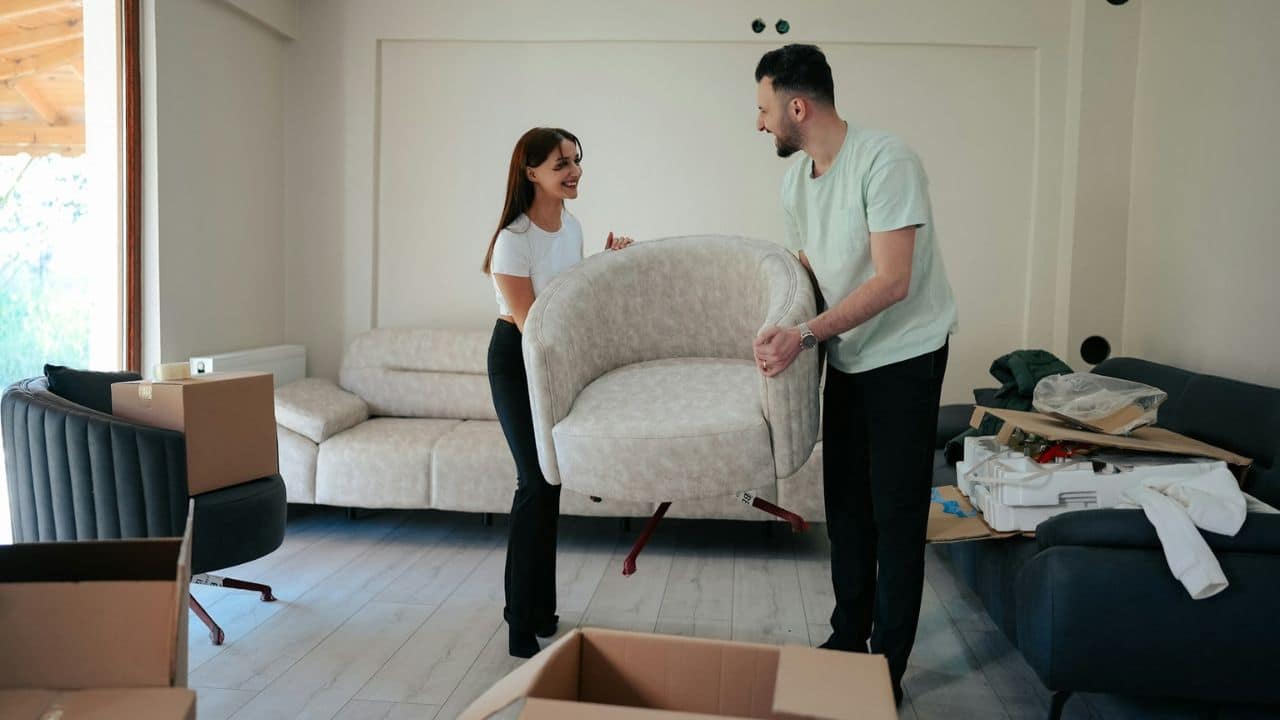 A man and woman smile at each other while carrying an armchair in a room with boxes.