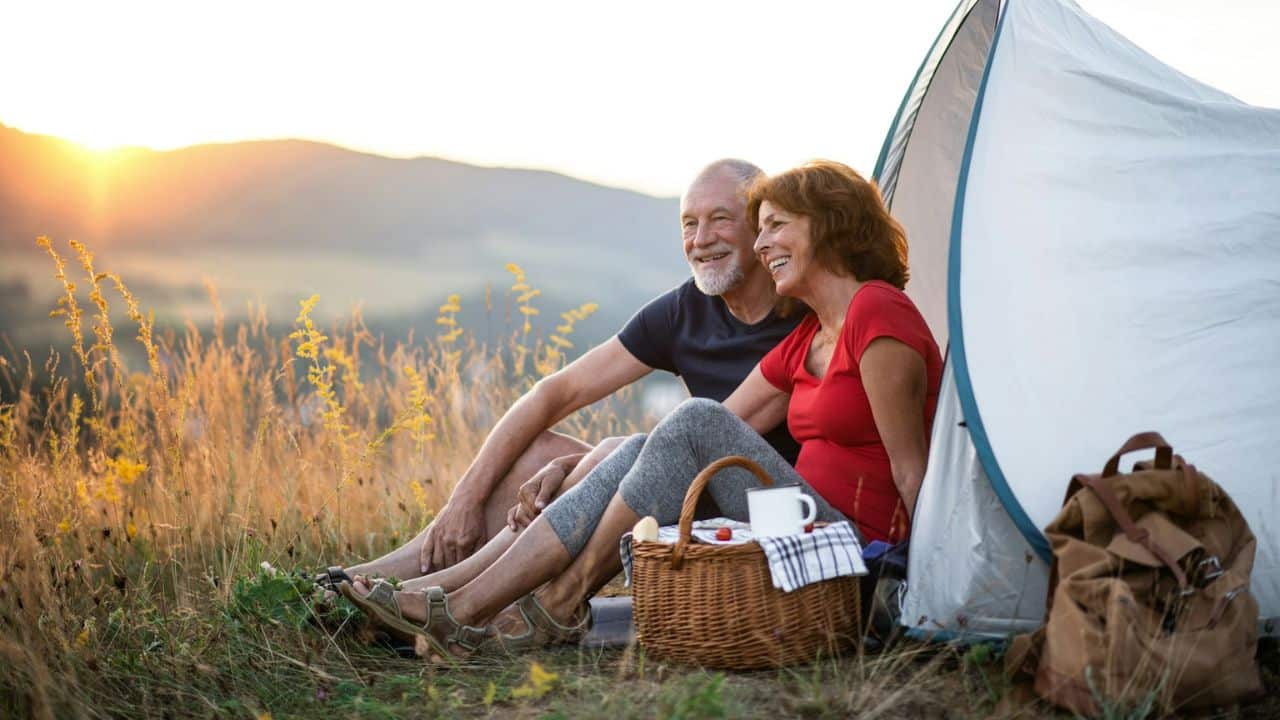 A smiling older couple sits beside a tent with a picnic basket, enjoying the sunset.