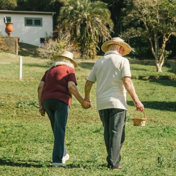 A senior couple, wearing straw hats, holds hands while walking across a grassy field.