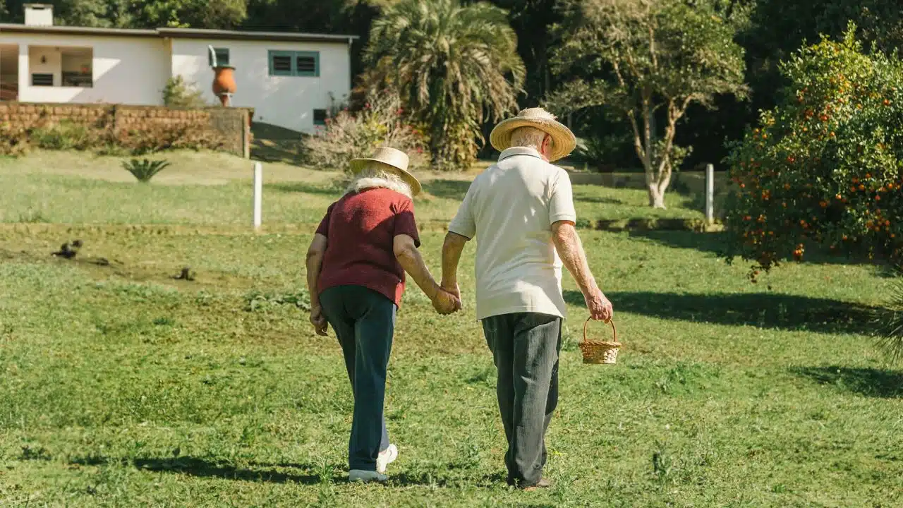 A senior couple, wearing straw hats, holds hands while walking across a grassy field.