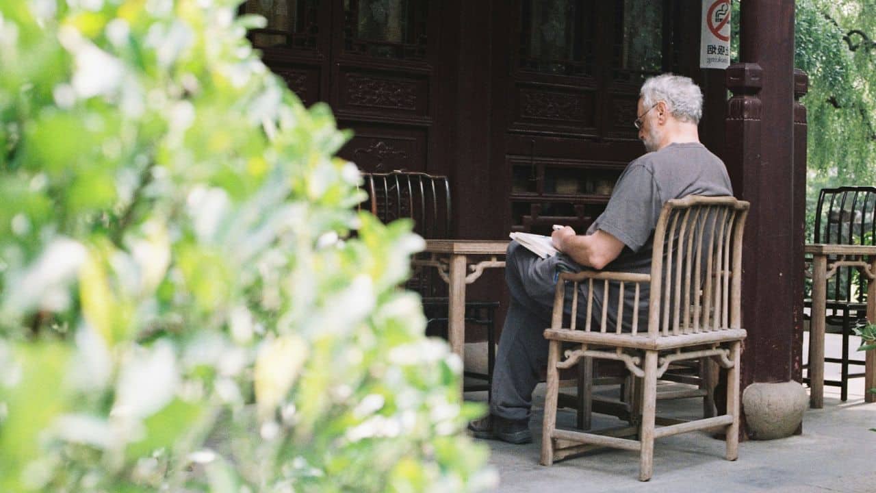 A man with gray hair and glasses sits on a wooden chair, writing in a book outdoors.