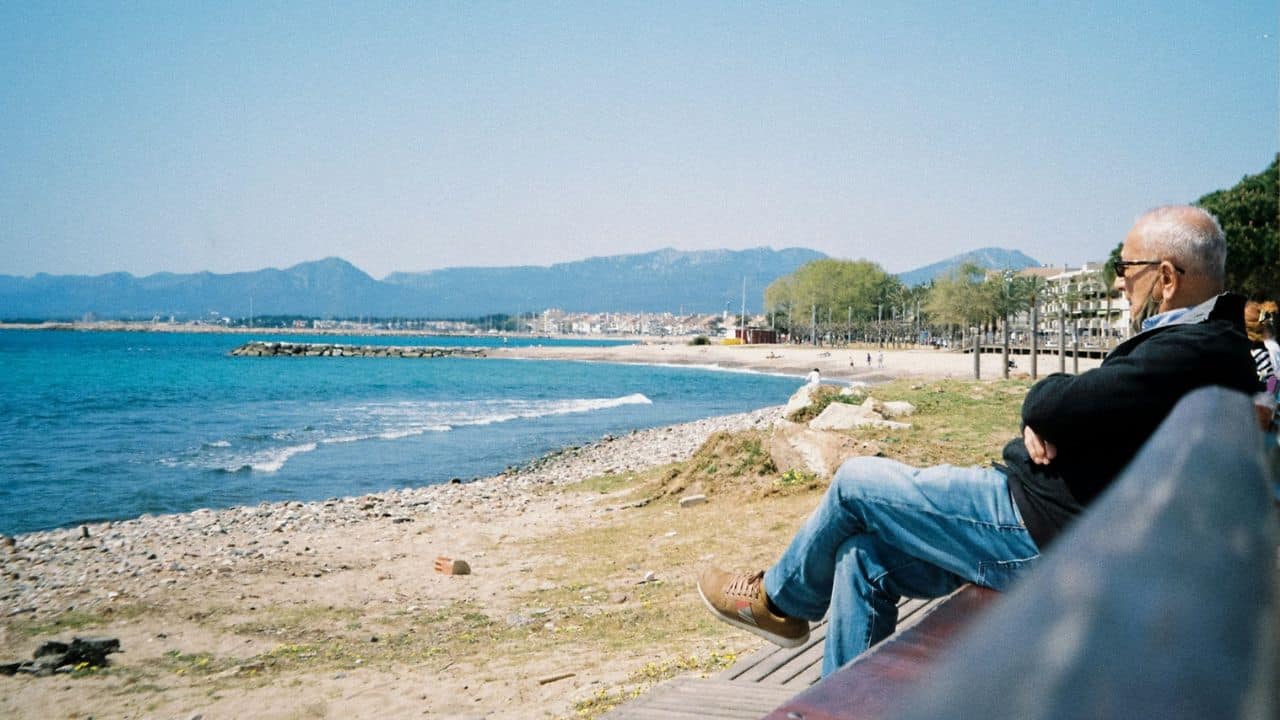 A man sits on a bench facing a beach with blue water and mountains in the distance.