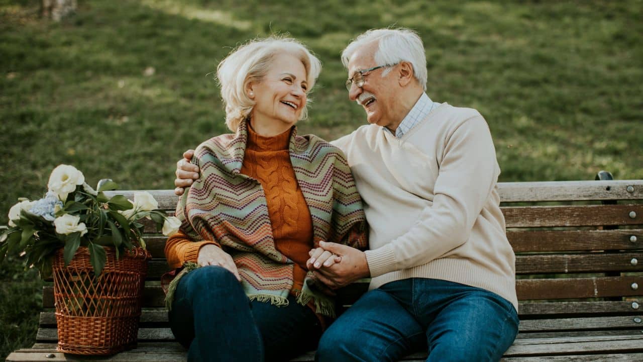 A smiling older couple sits on a park bench, holding hands, with a flower basket.