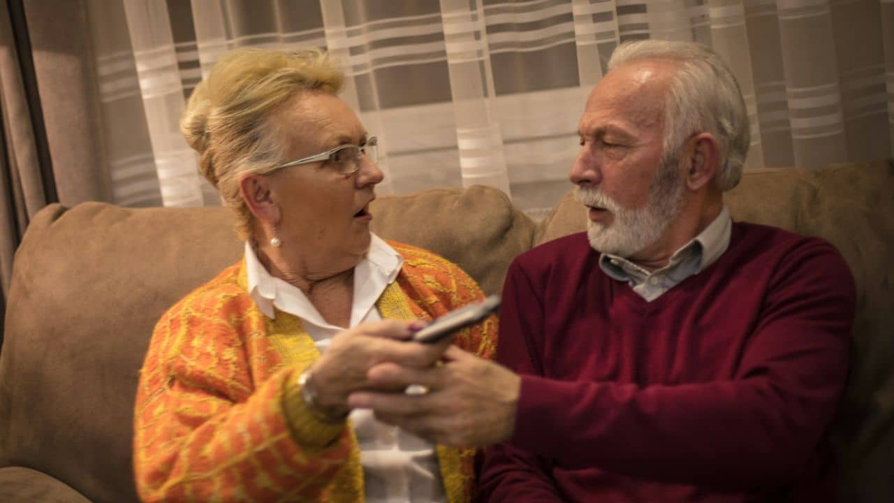 A senior woman in an orange cardigan argues with a man in a red sweater over a remote.