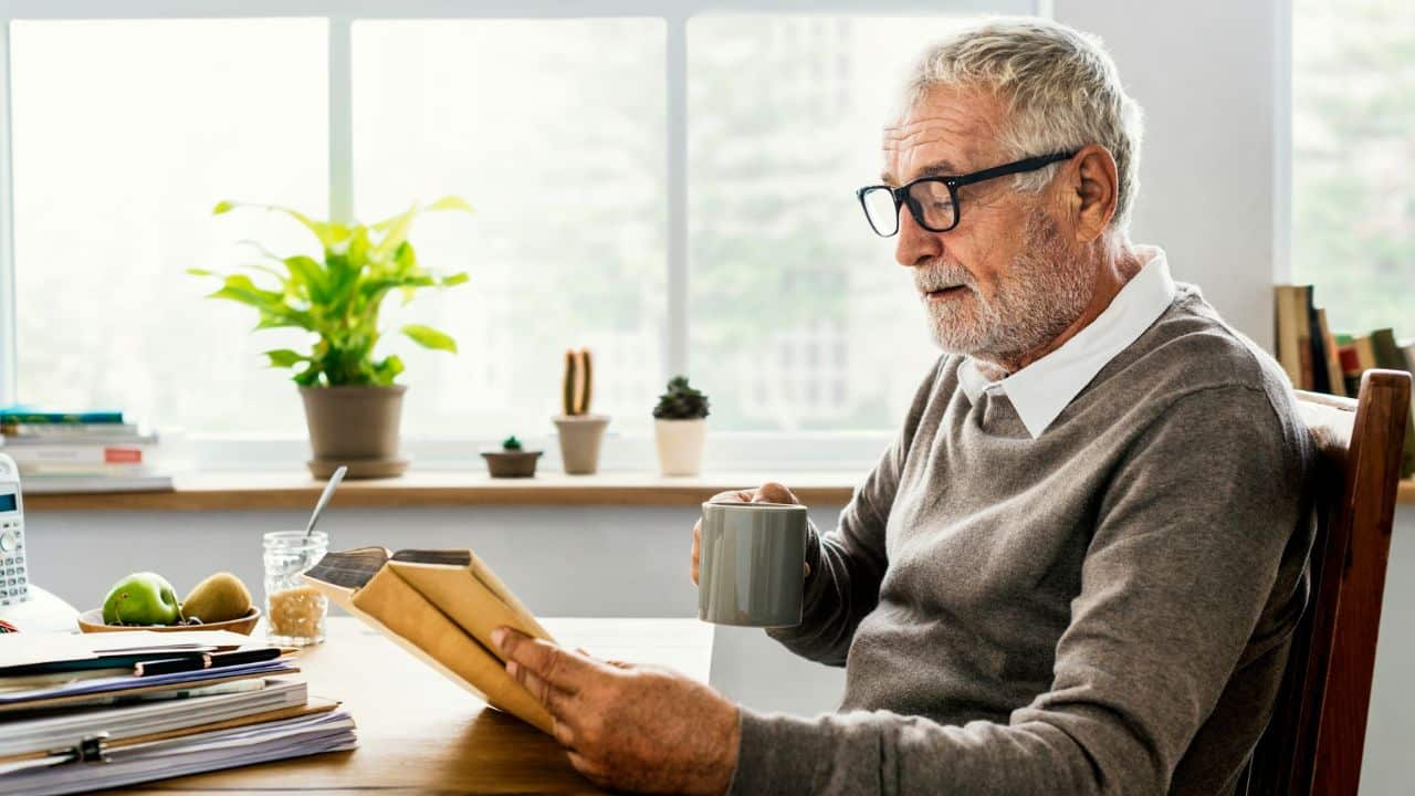 A bearded man with glasses holds a mug and reads a book at a table.