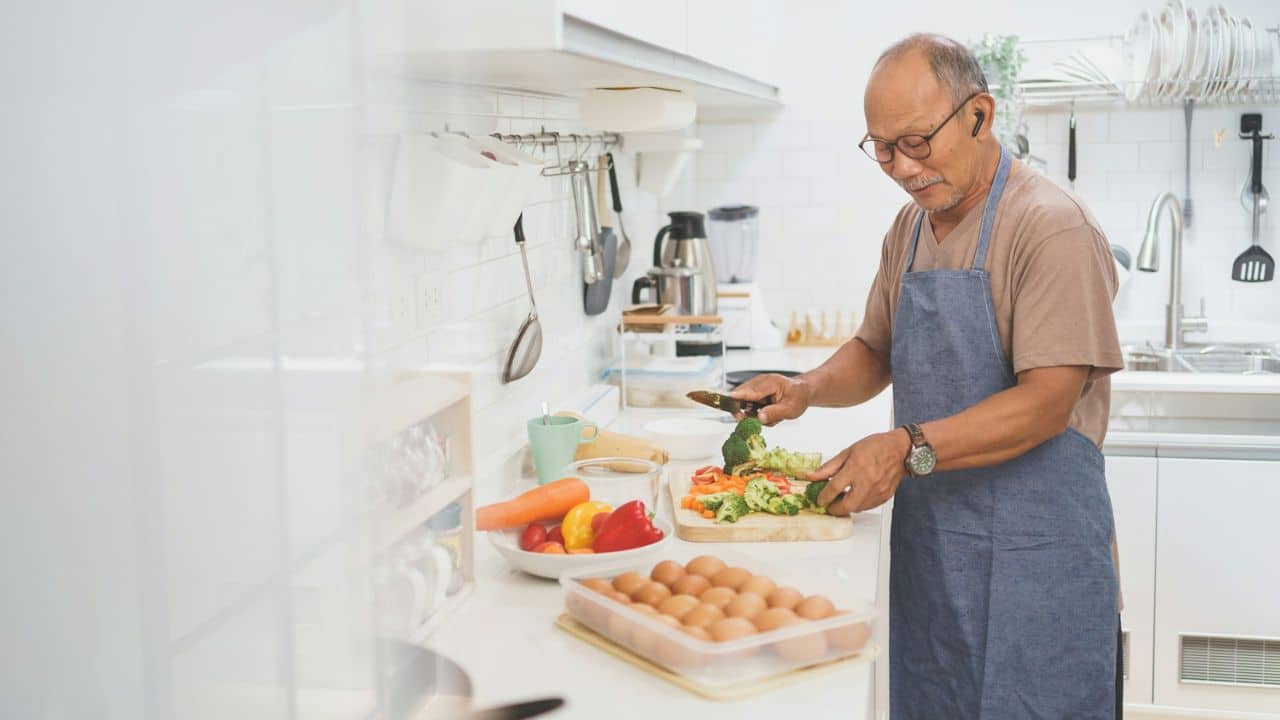 A man wearing glasses and an apron chops vegetables in a bright kitchen.
