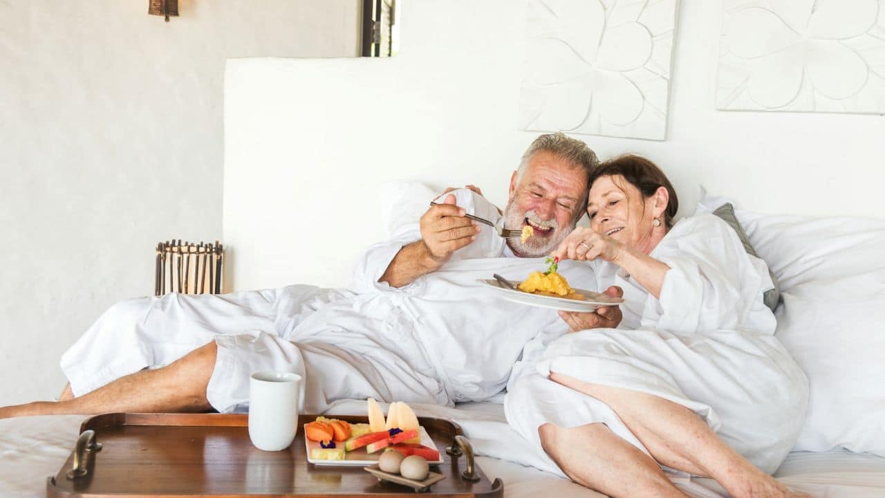 A happy elderly couple in white robes eats breakfast in bed, smiling and sharing food.