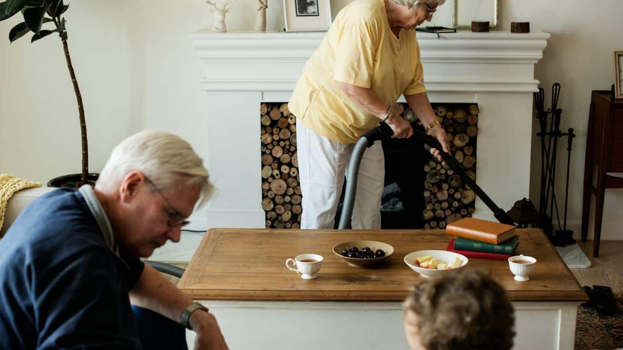 A woman vacuums near a fireplace while a man sits by a coffee table with snacks.