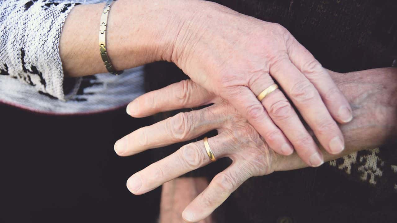 A close-up shows an older couple's hands with wedding rings, gently clasped together.