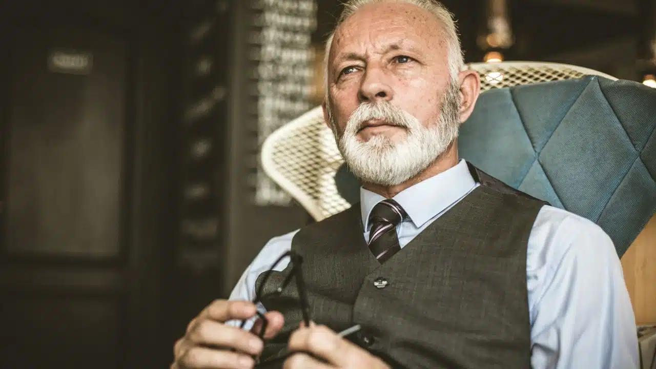 A distinguished older man with a white beard sits in a chair, holding his glasses.