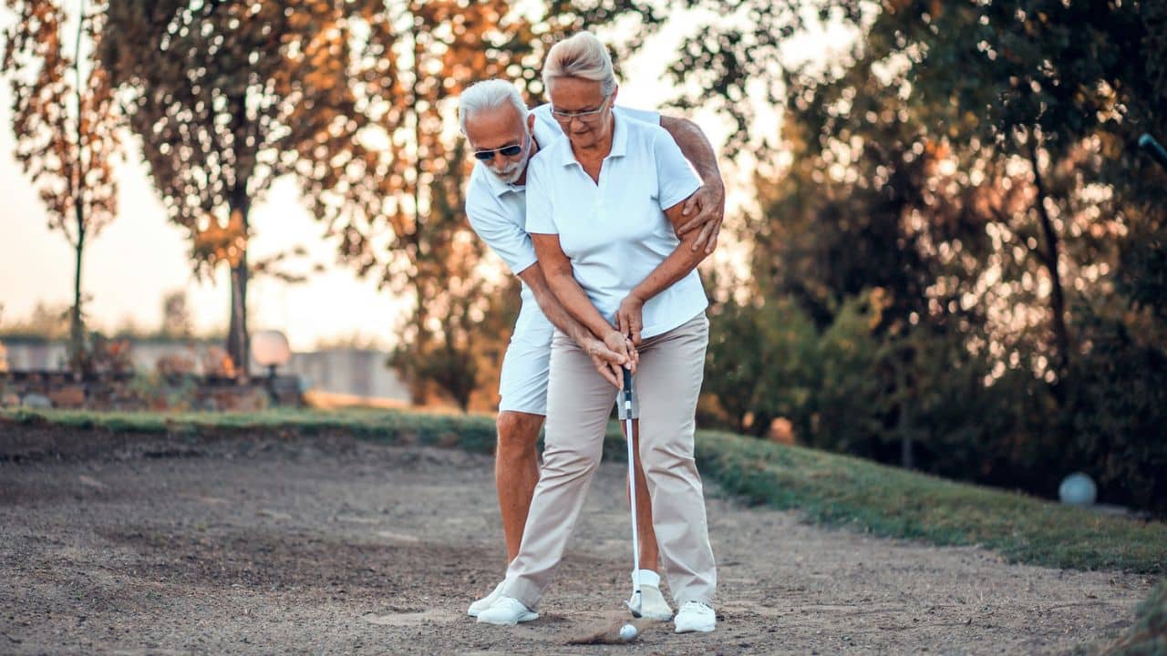 A man in a white polo shirt helps a woman in a white polo shirt swing a golf club.