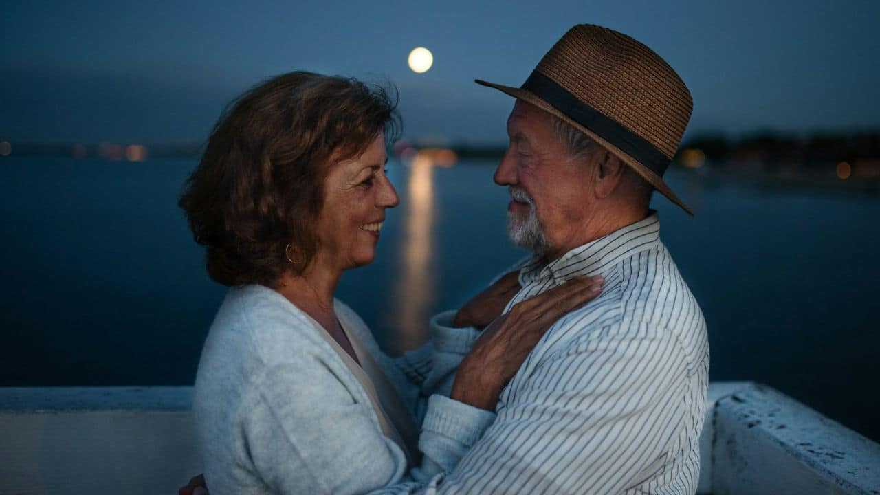 A smiling older couple gazes at each other under a full moon by the water.