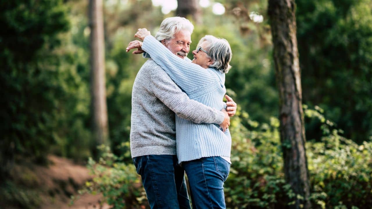 A smiling older couple with grey hair embraces outdoors, surrounded by trees.